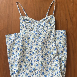 Blommig långklänning med snörning i ryggen - Säljer en söt långklänning med smala axelband och blått blommönster på vit botten. Klänningen har snörning i ryggen och en slits framtill för extra detalj. Perfekt för varma dagar och somriga tillfällen. 