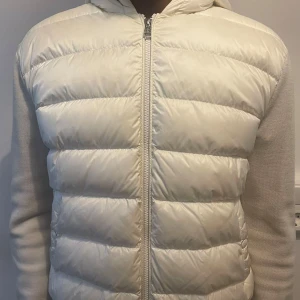 Vit pufferjacka med huva från Moncler - Säljer en stilren vit pufferjacka från Moncler med huva och dragkedja framtill. Jackan har quiltade partier på framsidan och släta stickade ärmar med Moncler-logga och röd/blå rand på ärmen. Perfekt för dig som gillar exklusiv streetwear. (Även öppen för byten)