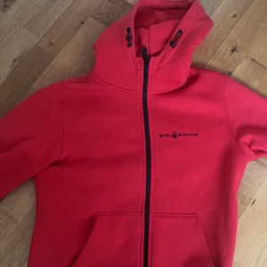 Röd hoodiejacka från Sail Racing - Röd hoodiejacka från Sail Racing med svart dragkedja och logga på bröstet. Jackan har huva med snörning och två fickor framtill. Perfekt för en sportig och avslappnad stil.