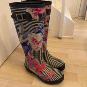 Randiga blommiga gummistövlar från Joules - Säljer ett par höga gummistövlar från Joules med svartvita ränder och stora blommor i rosa, lila och beige. Stövlarna har spänndetalj upptill och grov sula. Perfekta för regniga dagar.