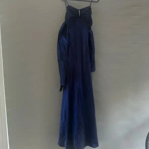 Marinblå långklänning med rosett och strassdetalj - Säljer en marinblå långklänning med smala axelband och stor rosett framtill, dekorerad med strass. Perfekt för dig som vill sticka ut på bal!. Passar s men även M