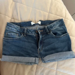 Lågmidjade Shorts (sydda från jeans till shorts) - Sytt själv! Snygga blå jeansshorts med klassisk femficksdesign och upprullad kant nedtill. Shortsen har bälteshällor och knappgylf. Perfekta för varma dagar och enkel att matcha med olika toppar.