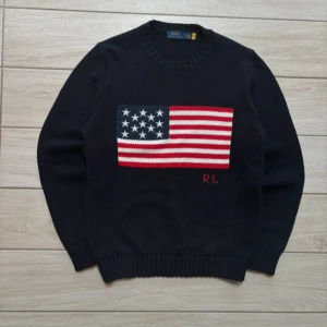 Mörkblå stickad tröja från Polo Ralph Lauren - Säljer en mörkblå stickad tröja från Polo Ralph Lauren med amerikansk flagga framtill och RL-broderi. Tröjan har rund halsringning och ribbade muddar vid ärmslut och nederkant. Tröjan är helt ny storlek M