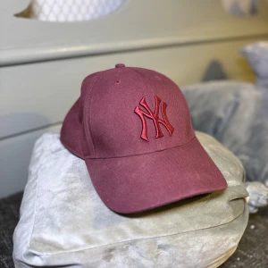 Vinröd New York Yankees keps - Snygg vinröd keps med New York Yankees-logga broderad framtill i matchande färg. Klassisk böjd skärm och justerbar passform. Perfekt accessoar för en sportig och trendig look.