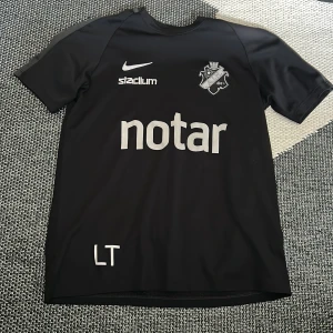 Svart AIK fotbollströja från Nike - Säljer en svart AIK fotbollströja från Nike med tryck från notar, Stadium och klubbmärke på bröstet. Tröjan har korta ärmar och är tillverkad i Dri-FIT material som andas. Sponsorer även på ryggen. Perfekt för träning eller match!
