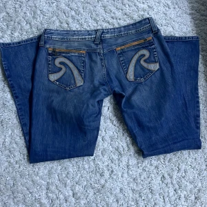 Blå Replay jeans med broderad bakficka - Säljer ett par blå jeans från Replay med snygga broderade detaljer och dragkedjor på bakfickorna. Klassisk femficksmodell med raka ben och orangea sömmar. Perfekta för dig som gillar jeans med unika detaljer.