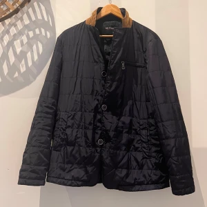 Svart quiltad jacka med bruna mocka detaljer.  - Snygg svart quiltad jacka med bruna mockadetaljer på krage och armbågar. Jackan har lång ärm, knäppning framtill med knappar och dragkedja vid bröstficka. Perfekt för höst och vår.