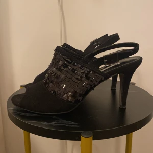 Svarta klackskor med paljettdetaljer - Säljer ett par svarta klackskor med öppen tå och snygga paljettdetaljer framtill. Skorna har en justerbar rem runt hälen och en elegant klack som ger extra höjd. Perfekta för att lyfta din outfit med lite extra glans.