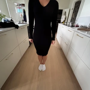 Svart långärmad klänning med v-ringning - En stilren svart klänning med lång ärm och v-ringad hals. Klänningen har en tight passform och går till knäna, vilket ger en elegant och enkel siluett. Perfekt för dig som gillar minimalistisk stil. Klänningen är ribb stickad.