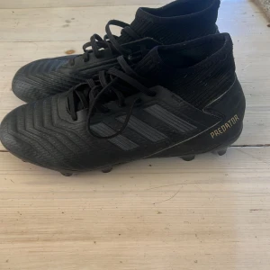 Svarta Adidas Predator fotbollsskor - Säljer ett par svarta Adidas Predator fotbollsskor med snörning och klassisk design. Skorna har en högre strumpliknande krage och tre svarta ränder på sidan. Perfekta för dig som vill ha bra grepp på planen. 