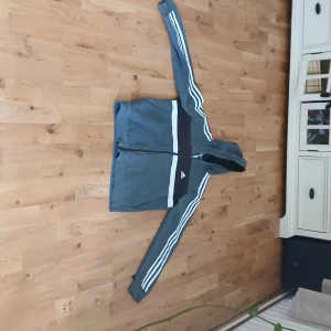En flis från Adidas  - Säljer en grå hoodie från Adidas med vit randdetalj på ärmarna och bröstet. Jackan har dragkedja framtill, huva och klassiska Adidas-loggan på bröstet. Perfekt för träning eller chill.