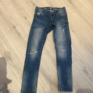 Blå jeansbyxor från LAB Industries - Säljer ett par blå jeansbyxor från LAB Industries, modell Travis Regular Fit. Byxorna har slitna detaljer och hål på framsidan för en trendig look. Klassisk femficksmodell med normal passform och raka ben.