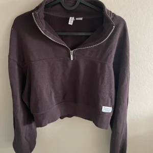 Brun croppad sweatshirt med dragkedja från H&M - Mörkbrun croppad sweatshirt från H&M med halv dragkedja framtill och hög krage. Tröjan har långa ärmar och en liten vit patch nertill. Perfekt för en avslappnad stil.
