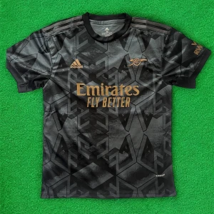 Svart Arsenal fotbollströja från Adidas - Säljer en svart Arsenal fotbollströja från Adidas med guldiga detaljer och loggor. Tröjan har ett geometriskt mönster och korta ärmar. Perfekt för dig som vill sticka ut på planen eller på läktaren.