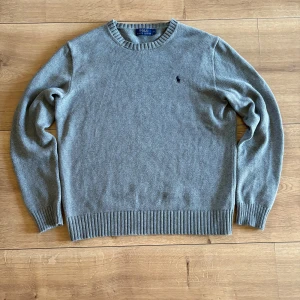 Grå stickad tröja från Polo Ralph Lauren - Klassisk grå stickad tröja från Polo Ralph Lauren med rund halsringning och broderad logga på bröstet. Tröjan har ribbade muddar vid ärmslut och nederkant. Perfekt för lager-på-lager och enkel att matcha.