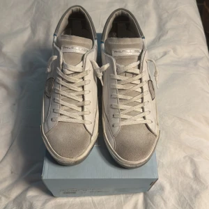 Vita Philippe Model sneakers med grå detaljer - Säljer ett par vita Philippe Model sneakers med grå/silvriga detaljer på tån och hälen. Skorna har snörning, rund tå och en platt sula. Märkets logga syns på sidan och bak på hälen. Perfekta för en stilren och avslappnad look.