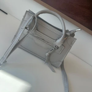 Ljusgrå mini axelväska med dragkedja - Säljer en ljusgrå mini axelväska med både handtag och lång axelrem. Väskan har en snygg dragkedjedetalj framtill och är tillverkad i syntetmaterial. Perfekt för dig som vill ha en stilren och smidig accessoar till vardagslooken.