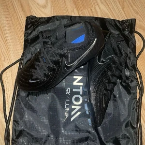 Svarta Nike fotbollsskor Phantom GX - Säljer ett par svarta Nike Phantom GX fotbollsskor med silverfärgade detaljer och blå insida. Skorna har snörning och är designade för bra grepp på planen. Perfekta för dig som vill ta spelet till nästa nivå.