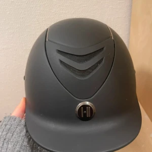 Svart ridhjälm från OneK - Säljer en stilren svart ridhjälm från OneK med ventilerande meshdetaljer framtill och diskret logotyp. Hjälmen har en klassisk form med liten skärm och mattsvart finish. Perfekt för ridning och tävling.