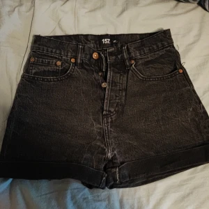 Svarta jeansshorts från 157 - Säljer ett par svarta jeansshorts från 157 i storlek XS. De är högmidjade med klassisk femficksdesign, uppvikta benslut och knappar framtill. Perfekta för sommaren och enkla att matcha med olika toppar.