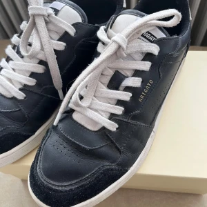 Svarta sneakers från Axel Arigato - Snygga svarta sneakers från Axel Arigato med vita detaljer och snörning. Skorna är i skinn med vit sula och har klassisk låg profil. Märkeslogga i guld på sidan och vit plös med svart logotyp. Perfekta för en clean och stilren look.
