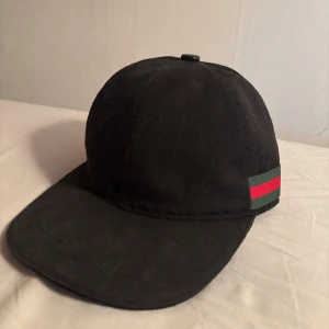 Svart Gucci canvas keps - Snygg svart keps från Gucci med klassisk röd och grön rand på sidan. Kepsen har böjd skärm och justerbar rem baktill. Materialet är bomull och designen är stilren med diskret Gucci-mönster över hela kepsen. Perfekt för dig som vill ha en lyxig streetstyle-accessoar. Kepsen är äkta och i gott skick hör av er vid frågor. Pris går att diskutera.