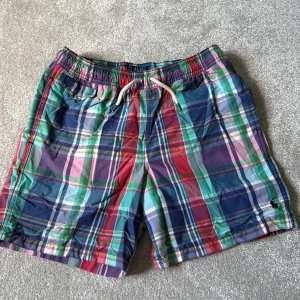 Rutig badshorts från Polo Ralph Lauren - Färgglada rutig badshorts från Polo Ralph Lauren med snörning i midjan och meshfoder. Shortsen har flera fickor och klassisk Polo-logga broderad på benet. Mix av blå, röd, grön, rosa och lila toner. Perfekt för sommaren och strandhäng.