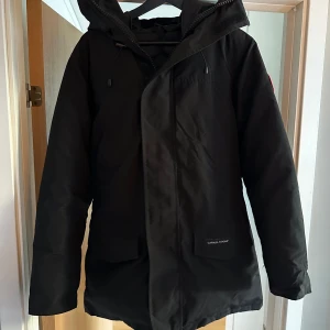 Svart dunjacka från Canada Goose - Säljer en svart dunjacka från Canada Goose med huva och klassisk logga på ärmen. Jackan har dragkedja, stora fickor framtill och justerbar dragsko i midjan. Perfekt för kalla vinterdagar och riktigt snygg streetstil.