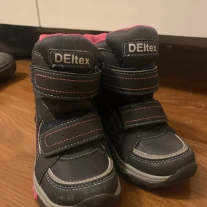 Svarta vinterboots från DELtex med rosa detaljer - Säljer ett par svarta vinterboots från DELtex med rosa insida och rosa detaljer på sulan. Skorna har två breda kardborreband för enkel påtagning och en snöflinga på sidan. Grov sula och slitstarka sömmar, perfekta för kalla dagar.