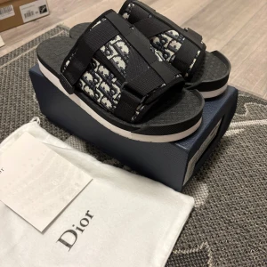 Svarta sandaler från Dior med monogram - Snygga sandaler från Dior med svart och vit monogrammönstrad textil, breda remmar och präglad sula med Dior-logga. Sulan är svart med vit kant och remmarna har justerbara detaljer. Perfekta för en trendig och avslappnad look.