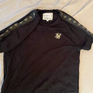 Svart t-shirt från SikSilk med logga - Svart t-shirt från SikSilk med guldfärgad broderad logga på bröstet och tryckta loggor längs ärmarna. Klassisk rund halsringning och korta ärmar. Storlek M