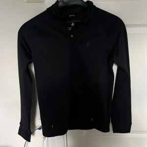 Svart sporttröja från Cross Sportswear - Svart långärmad sporttröja från Cross Sportswear med half zip framtill. Tröjan har en hög krage och är tillverkad i ett stretchigt syntetmaterial som passar perfekt för träning eller utomhusaktiviteter. Enkel och stilren design utan mönster.