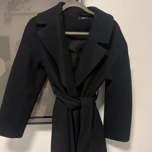 Lång Svart kappa med bälte från Gina Tricot - Stilren Lång svart kappa från Gina Tricot med bred krage och knytskärp i midjan. Välanvänd men i otroligt bra skick! XS men är oversized i storleken!