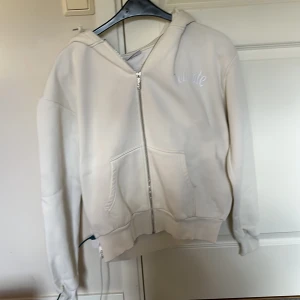 Vit hoodie med dragkedja från Elevate - Säljer en stilren vit hoodie från Elevate i storlek XS. Tröjan har huva med snörning, hel dragkedja framtill och två stora fickor. Perfekt för dig som gillar en enkel och clean look. Materialet är mjukt och skönt, troligtvis bomull eller polyester.