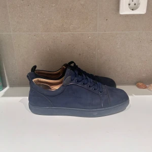 Mörkblå Louboutin - Snygga mörkblå sneakers i mocka från Christian Louboutin med klassisk röd sula. Skorna har rund tå, platt sula och snörning. Broderad logga baktill och lyxig känsla både på utsidan och insidan. Box finns 
