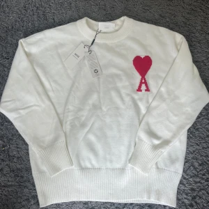Vit stickad tröja från Ami Paris - Vit stickad tröja från Ami Paris med rund hals och ribbade muddar. På bröstet finns ett rött hjärta med bokstaven A. Tillverkad i ekologisk bomull, perfekt för dig som gillar stilrena och trendiga plagg med en twist.