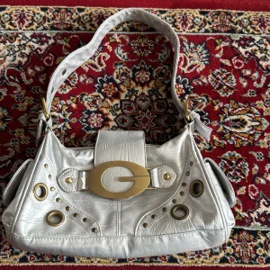 Silvrig axelväska med stora metalldetaljer - Cool silvrig axelväska som liknar Guess med stort G-spänne i guldmetall framtill. Väskan har bred axelrem, dragkedja och flera runda metalldetaljer 