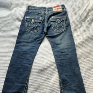 True Religion blå jeans med kontrastsömmar - Säljer ett par blå jeans från True Religion med tydliga vita kontrastsömmar och ikoniska ficklock bak med stora knappar. Klassisk femficksmodell och snygg tvättad look. Märkespatch bak i midjan och detaljerade nitar framtill. Perfekta för dig som gillar street och premium denim.
