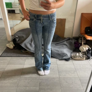 Blå bootcut jeans med pressveck - Säljer ett par ljusblå bootcut jeans med markerade pressveck framtill. Jeansen har låg midja, klassiska fem fickor och dubbla knappar i linningen. Snygg tvätt och skön passform, perfekta till sneakers eller boots.