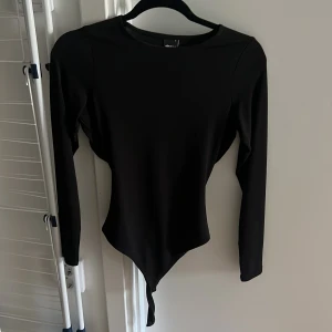 Svart långärmad bodysuit med cut-out - Svart bodysuit från Ginatricot med lång ärm och cool cut-out detalj i ryggen. Tight passform som framhäver midjan och ger en edgy vibe. Materialet är mjukt och stretchigt, perfekt för att skapa en snygg siluett. Rund halsringning och axelparti med markerad söm.