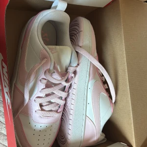 Nike court borough low recraft (GS) - Snygga Nike sneakers i vitt och ljusrosa med klassisk swoosh på sidan. Skorna har snörning, rund tå och platt sula. Ovandelen är i syntet och skinn, med perforerade detaljer framtill för extra ventilation. Perfekta för dig som gillar en clean och sportig look.