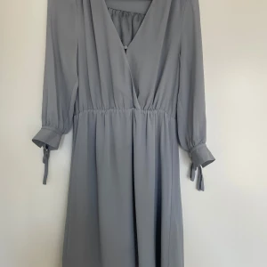 Grå omlottklänning från H&M - Supersnygg grå klänning från H&M med omlott-v-ringning och långa ärmar med knyt vid mudden. Klänningen har en lätt rynkad midja som ger en skön passform och ett flowy fall. Perfekt för dig som gillar stilrena och enkla plagg med en twist.