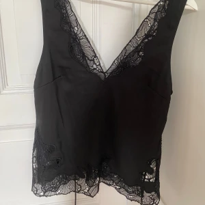 Svart linnetopp med spetsdetaljer - Säljer denna svarta satintopp med spetsdetaljer. Den är öppen i ryggen med en knytning. Perfekt till fest eller om man vill vara lite mer uppklädd!😍😍använd max 2 gånger och nypris 400 kr