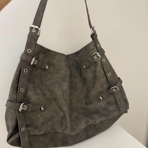 Grå axelväska i mocka från Monki - Snygg grå axelväska från Monki i mocka-look med spänndetaljer och silvriga metalldelar. Väskan har innerfack med dragkedja och en bred axelrem. Perfekt om man vill få plats med mycket t.e.x  skoldator.          (På andra bilden har jag fyllt med lite grejer). Väskan är utöver det i bra skick.