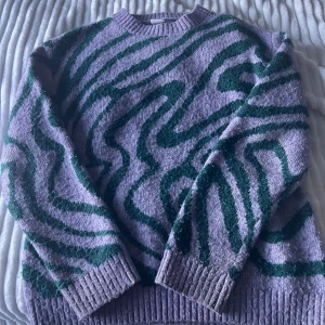 Lila stickad tröja med grönt mönster Lindex - Säljer en lila stickad tröja från Lindex med gröna vågiga mönster. Tröjan har rund halsringning, ribbade muddar och är långärmad. Perfekt för dig som gillar färg och unika mönster!