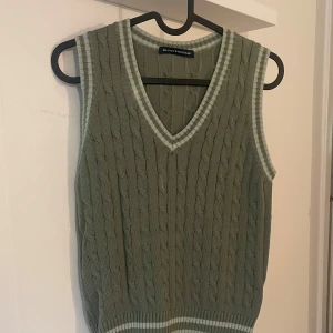 Olivgrön stickad väst Brandy Melville - Snygg olivgrön stickad väst från Brandy Melville med klassiskt kabelstickat mönster. V-ringad hals och ribbade, randiga detaljer i ljusgrönt och vitt vid hals, ärm och nederkant. 