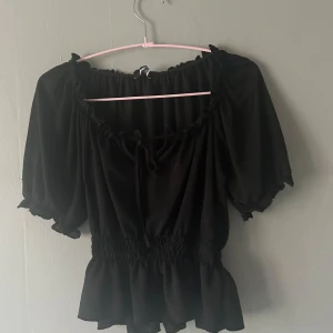 Svart blus med volang från Gina Tricot - Supersöt svart blus från Gina Tricot i storlek XS. Modellen har korta puffärmar, smock i midjan och volangkant nertill. Urringningen är dekorerad med en liten knytning och volangdetaljer. Tunt och lätt material som känns luftigt.