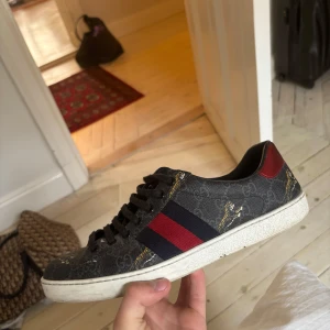 Gucci ace  - Snygga Gucci sneakers i mörkgrått med klassiskt GG-mönster, röda och blå ränder på sidan och detaljer med guldiga tigrar. Skon har svarta skosnören, vit sula och röd hälkappa. Perfekt för dig som vill sticka ut med lyxig streetstyle.