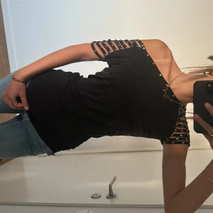 Svart offshoulder topp med nitar - Cool svart offshoulder topp med unika banddetaljer och silvriga nitar längs ärmar och halsringning. Ärmarna är korta och består av flera band som ger en edgy vibe. Perfekt för dig som vill sticka ut med en trendig och snygg look.