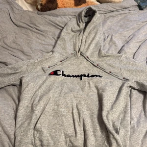 Grå hoodie från Champion, Small - Säljer en grå hoodie från Champion i storlek Small. Tröjan har broderad Champion-logga på bröstet och snören med logotypdetaljer. Mjuk insida och klassisk känguruficka framtill. Perfekt för chill dagar eller när du vill ha en sportig look.